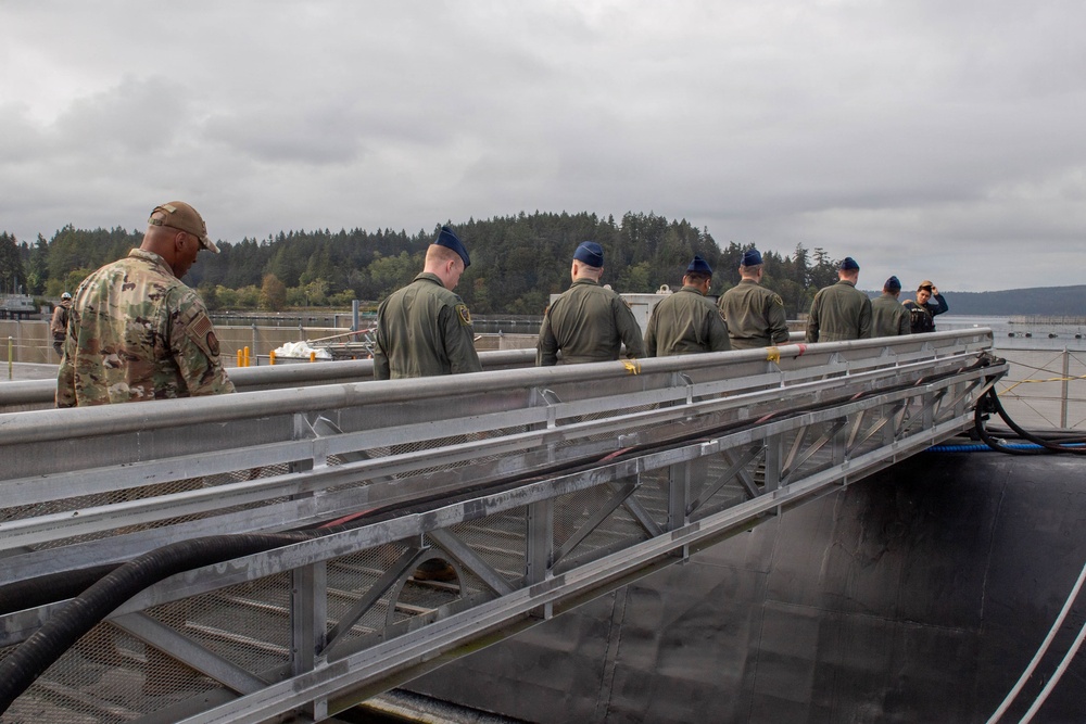 A group of military personnel in uniform are walking along a gangway or brow on what appears to be the deck of a submarine at a naval facility. One individual on the left wears Army OCP (camouflage) uniform while the majority wear olive/sage green flight suits, suggesting an Air Force contingent visiting a Navy submarine. The setting appears to be a Pacific Northwest naval base, indicated by the forested shoreline and overcast sky.