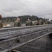 A group of military personnel in uniform are walking along a gangway or brow on what appears to be the deck of a submarine at a naval facility. One individual on the left wears Army OCP (camouflage) uniform while the majority wear olive/sage green flight suits, suggesting an Air Force contingent visiting a Navy submarine. The setting appears to be a Pacific Northwest naval base, indicated by the forested shoreline and overcast sky.