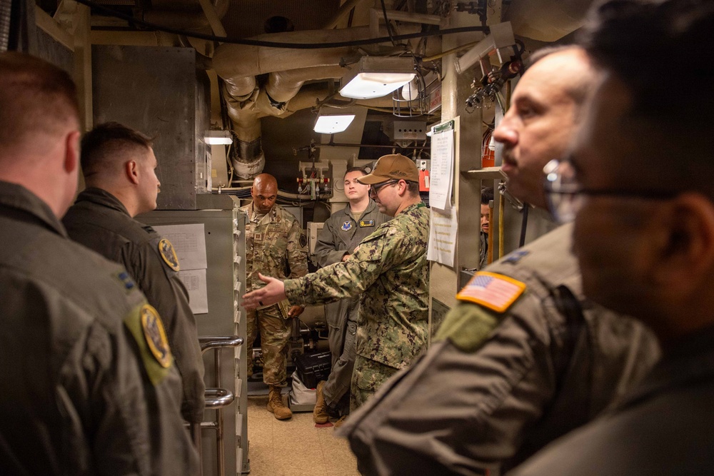A group of multi-branch U.S. military personnel are gathered in a narrow, equipment-filled corridor or compartment, likely aboard a naval vessel or military facility. A Navy sailor in digital camouflage (NWU) appears to be briefing or explaining something to personnel from other branches, including Army and Air Force members. The setting suggests an inter-service orientation, tour, or training exercise.