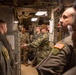 A group of multi-branch U.S. military personnel are gathered in a narrow, equipment-filled corridor or compartment, likely aboard a naval vessel or military facility. A Navy sailor in digital camouflage (NWU) appears to be briefing or explaining something to personnel from other branches, including Army and Air Force members. The setting suggests an inter-service orientation, tour, or training exercise.