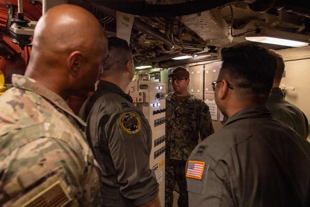 A group of military personnel in various uniforms are gathered in what appears to be a confined interior space of a naval vessel or military facility. One individual in Navy Working Uniform (NWU) camouflage pattern appears to be briefing or instructing the others near an equipment panel. The group includes personnel wearing Army combat uniforms, flight suits with patches, and Navy uniforms.