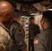 A group of military personnel in various uniforms are gathered in what appears to be a confined interior space of a naval vessel or military facility. One individual in Navy Working Uniform (NWU) camouflage pattern appears to be briefing or instructing the others near an equipment panel. The group includes personnel wearing Army combat uniforms, flight suits with patches, and Navy uniforms.