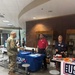 An indoor event featuring information booths for the DVA (Department of Veterans Affairs) and USO organizations. A military service member in Army Combat Uniform (ACU) is interacting with representatives at the DVA table. The setting appears to be a government or military building lobby with display cases and brick accent walls.