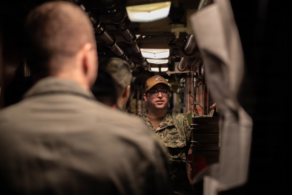 A U.S. Navy sailor wearing camouflage utilities and a tan cap is speaking to a group of personnel in what appears to be a confined interior space, likely aboard a naval vessel. The sailor appears to be conducting a briefing or tour in a narrow corridor with visible pipes and equipment overhead. Other military personnel are visible from behind in the foreground.