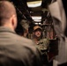 A U.S. Navy sailor wearing camouflage utilities and a tan cap is speaking to a group of personnel in what appears to be a confined interior space, likely aboard a naval vessel. The sailor appears to be conducting a briefing or tour in a narrow corridor with visible pipes and equipment overhead. Other military personnel are visible from behind in the foreground.