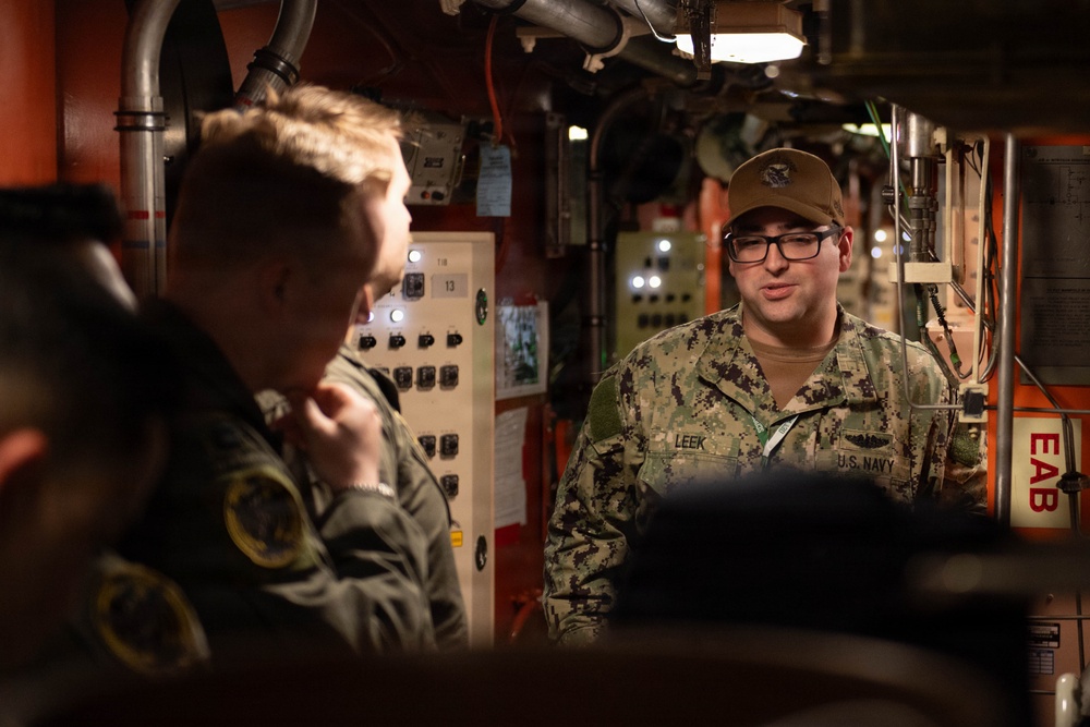 A U.S. Navy sailor identified by the name tape 'LEEK' is briefing or speaking to a group of individuals in what appears to be the interior of a naval vessel, likely a submarine or ship's compartment. The sailor is wearing Navy Working Uniform (NWU Type III) with a tan cover and glasses. The setting features orange-painted bulkheads, control panels, and piping typical of a military vessel interior.