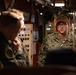 A U.S. Navy sailor identified by the name tape 'LEEK' is briefing or speaking to a group of individuals in what appears to be the interior of a naval vessel, likely a submarine or ship's compartment. The sailor is wearing Navy Working Uniform (NWU Type III) with a tan cover and glasses. The setting features orange-painted bulkheads, control panels, and piping typical of a military vessel interior.