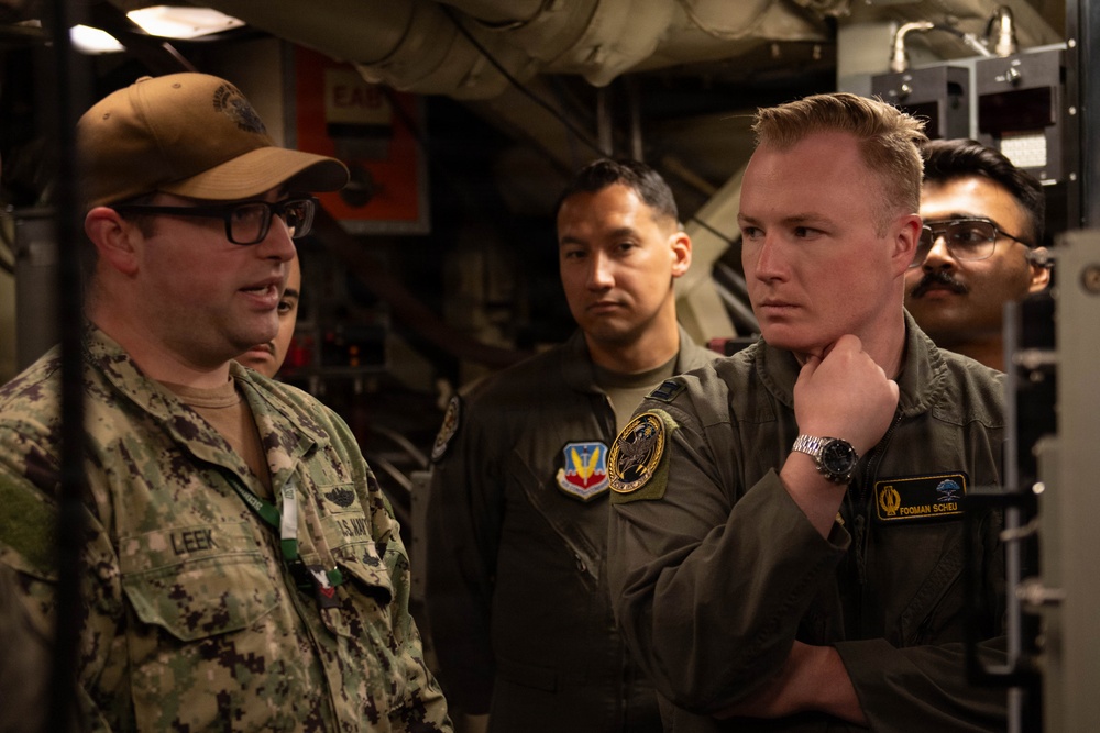 The image shows a group of military personnel from different branches engaged in a discussion inside what appears to be a naval vessel or military facility. A U.S. Navy sailor identified by the name tape 'LEEK' wearing digital camouflage and a tan cap appears to be briefing or explaining something to others. Among the group is a person in an olive flight suit with the name tape 'FORMAN SCHEU' and another individual wearing an Air Force flight suit with a squadron patch.