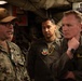 The image shows a group of military personnel from different branches engaged in a discussion inside what appears to be a naval vessel or military facility. A U.S. Navy sailor identified by the name tape 'LEEK' wearing digital camouflage and a tan cap appears to be briefing or explaining something to others. Among the group is a person in an olive flight suit with the name tape 'FORMAN SCHEU' and another individual wearing an Air Force flight suit with a squadron patch.