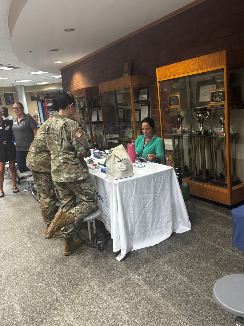 A military information or resource fair is taking place inside what appears to be a military building or installation. A soldier in Army Combat Uniform (ACU/OCP camouflage) is seated at a table speaking with a civilian woman in a green top who is representing an organization. Display cases with trophies and awards are visible in the background.