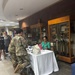 A military information or resource fair is taking place inside what appears to be a military building or installation. A soldier in Army Combat Uniform (ACU/OCP camouflage) is seated at a table speaking with a civilian woman in a green top who is representing an organization. Display cases with trophies and awards are visible in the background.
