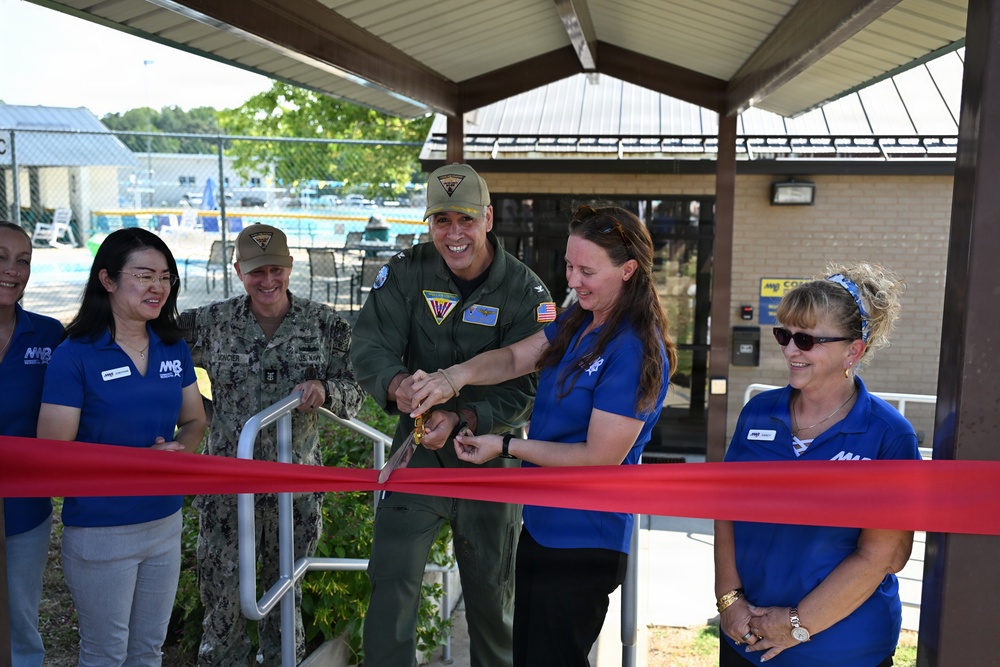 A ribbon-cutting ceremony is taking place at what appears to be a military recreational facility, likely a pool area. A military officer in a flight suit and a woman in a blue polo shirt are jointly cutting a red ribbon with golden scissors. Several other attendees, including a Navy personnel member in camouflage uniform and staff wearing blue 'MWR' (Morale, Welfare, and Recreation) polo shirts, are present and smiling.