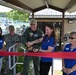A ribbon-cutting ceremony is taking place at what appears to be a military recreational facility, likely a pool area. A military officer in a flight suit and a woman in a blue polo shirt are jointly cutting a red ribbon with golden scissors. Several other attendees, including a Navy personnel member in camouflage uniform and staff wearing blue 'MWR' (Morale, Welfare, and Recreation) polo shirts, are present and smiling.