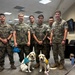 Six U.S. Marines in MARPAT camouflage uniforms pose indoors with two yellow Labrador Retrievers wearing service/therapy dog vests. The dogs appear to be service dog candidates or therapy dogs, identifiable by their colored vests (teal/blue and yellow). The setting appears to be an indoor military facility or office space.