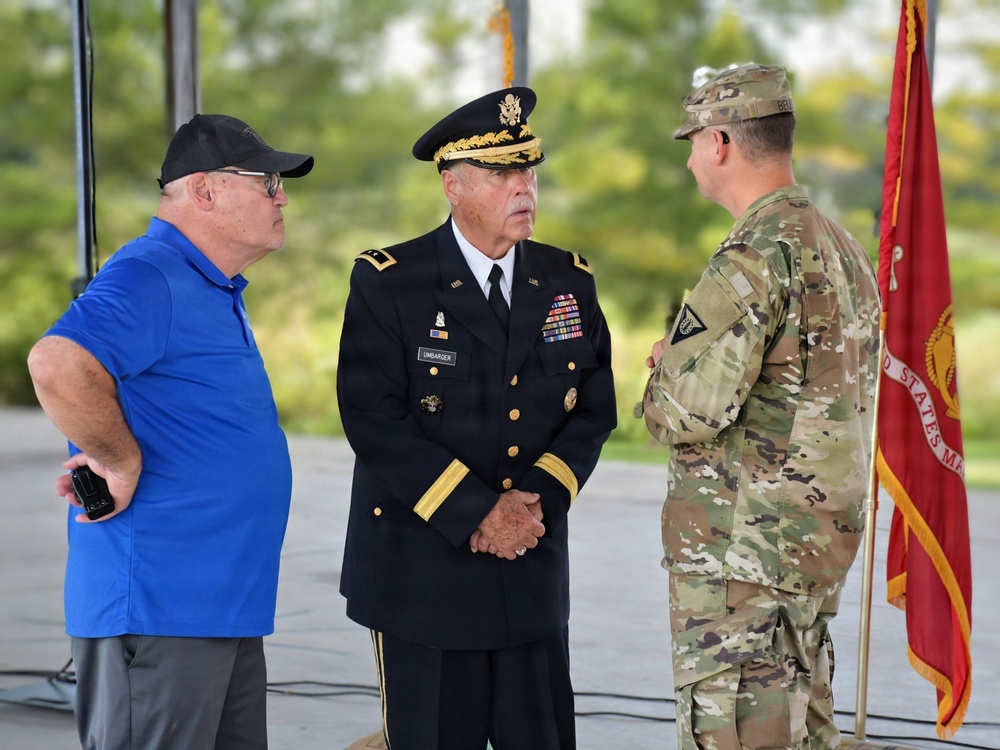 Three individuals are engaged in conversation at what appears to be an outdoor military ceremony or event. The central figure is a senior U.S. Army officer in dress uniform, flanked by a civilian in a blue polo shirt and another soldier in Army Combat Uniform (ACU/OCP camouflage). A red U.S. Army guidon or organizational flag is visible in the background.