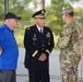 Three individuals are engaged in conversation at what appears to be an outdoor military ceremony or event. The central figure is a senior U.S. Army officer in dress uniform, flanked by a civilian in a blue polo shirt and another soldier in Army Combat Uniform (ACU/OCP camouflage). A red U.S. Army guidon or organizational flag is visible in the background.