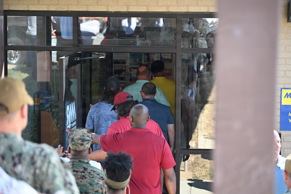 A crowd of people is entering or queuing to enter a building through glass double doors. At least one person in military camouflage uniform is visible among the civilians. The building appears to be a retail or service facility based on visible signage.
