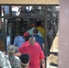 A crowd of people is entering or queuing to enter a building through glass double doors. At least one person in military camouflage uniform is visible among the civilians. The building appears to be a retail or service facility based on visible signage.