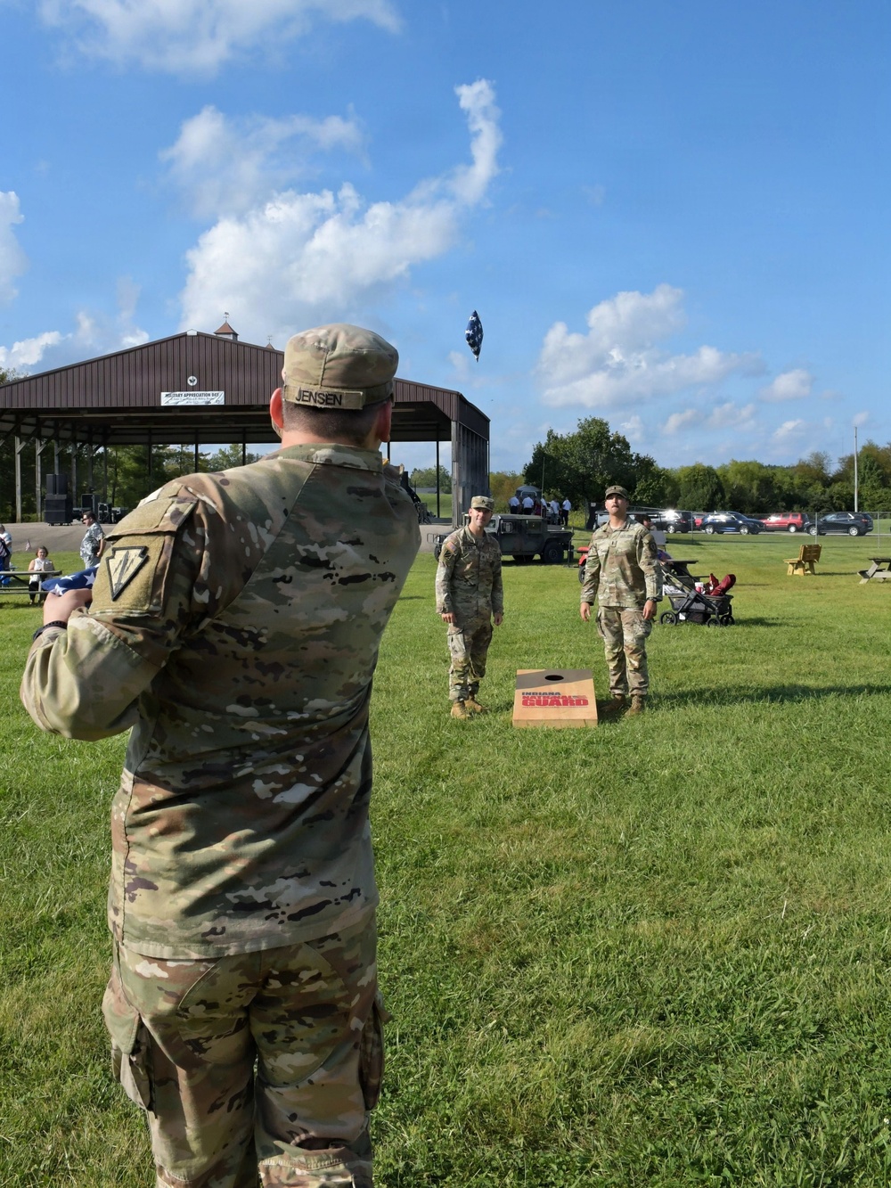 Three soldiers in U.S. Army OCP (Operational Camouflage Pattern) uniforms are playing cornhole at an outdoor military appreciation event. The soldier in the foreground, identified by name tape as 'JENSEN,' has just tossed a bean bag toward an Indiana National Guard-branded cornhole board. The event appears to be a casual outdoor gathering on a sunny day with a pavilion visible in the background.