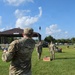 Three soldiers in U.S. Army OCP (Operational Camouflage Pattern) uniforms are playing cornhole at an outdoor military appreciation event. The soldier in the foreground, identified by name tape as 'JENSEN,' has just tossed a bean bag toward an Indiana National Guard-branded cornhole board. The event appears to be a casual outdoor gathering on a sunny day with a pavilion visible in the background.