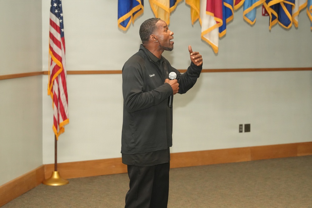 A man named 'Kevin' (as visible on his jacket) is speaking into a microphone in what appears to be a military or government facility. He is gesturing expressively with his right hand raised, suggesting an engaging presentation or motivational speech. Multiple military and organizational flags are displayed on the wall behind him, along with a U.S. flag on a stand.