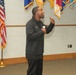 A man named 'Kevin' (as visible on his jacket) is speaking into a microphone in what appears to be a military or government facility. He is gesturing expressively with his right hand raised, suggesting an engaging presentation or motivational speech. Multiple military and organizational flags are displayed on the wall behind him, along with a U.S. flag on a stand.