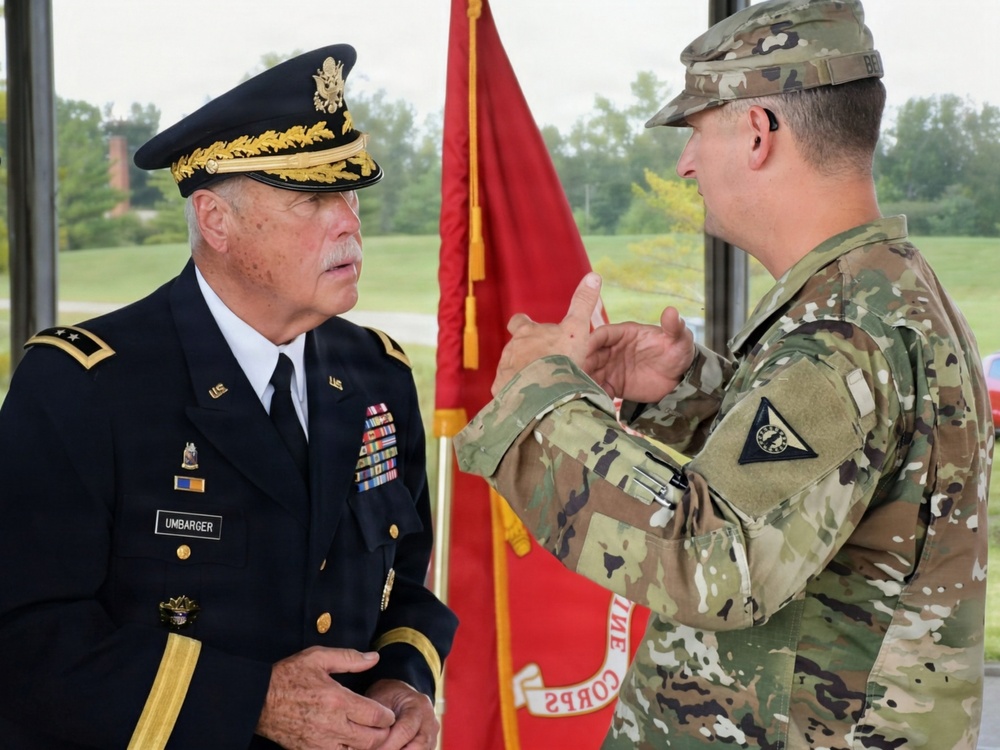 Two U.S. Army soldiers are engaged in conversation at what appears to be a military ceremony or formal event. The officer on the left wears a dress uniform with general officer rank insignia and a name tag reading 'UMBARGER', while the soldier on the right wears an OCP (Operational Camouflage Pattern) combat uniform. A red ceremonial flag/guidon is visible in the background.
