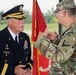 Two U.S. Army soldiers are engaged in conversation at what appears to be a military ceremony or formal event. The officer on the left wears a dress uniform with general officer rank insignia and a name tag reading 'UMBARGER', while the soldier on the right wears an OCP (Operational Camouflage Pattern) combat uniform. A red ceremonial flag/guidon is visible in the background.