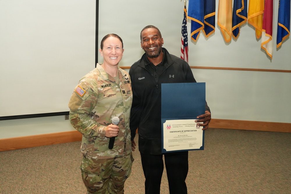 A U.S. Army soldier in OCP camouflage uniform, identified by name tape as 'McAuley,' stands alongside a civilian man named 'Kevin' who is holding a Certificate of Appreciation in a blue folder. The ceremony appears to take place in a military briefing room with multiple military unit flags displayed in the background. The soldier is holding a microphone, suggesting she was presiding over or announcing the award presentation.