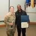 A U.S. Army soldier in OCP camouflage uniform, identified by name tape as 'McAuley,' stands alongside a civilian man named 'Kevin' who is holding a Certificate of Appreciation in a blue folder. The ceremony appears to take place in a military briefing room with multiple military unit flags displayed in the background. The soldier is holding a microphone, suggesting she was presiding over or announcing the award presentation.
