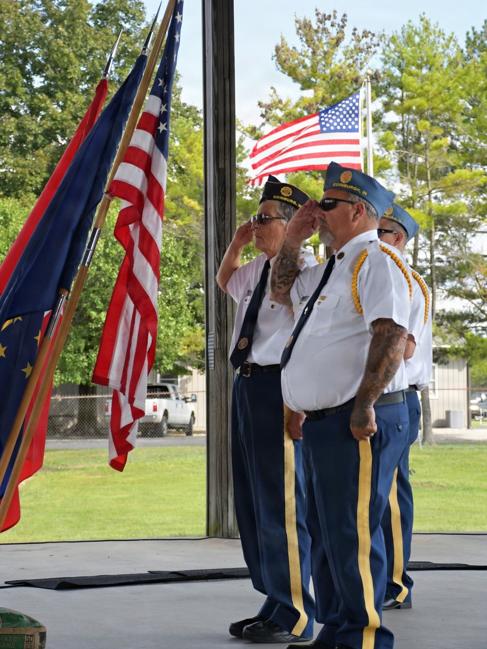Three members of an American Legion post from Edinburgh, Indiana are performing a salute during a color guard ceremony. They are dressed in white shirts with blue trousers featuring yellow stripes, wearing garrison caps. Multiple flags including the U.S. flag and what appears to be the Indiana state flag are displayed, with an additional U.S. flag visible on a flagpole outside.