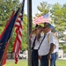 Three members of an American Legion post from Edinburgh, Indiana are performing a salute during a color guard ceremony. They are dressed in white shirts with blue trousers featuring yellow stripes, wearing garrison caps. Multiple flags including the U.S. flag and what appears to be the Indiana state flag are displayed, with an additional U.S. flag visible on a flagpole outside.