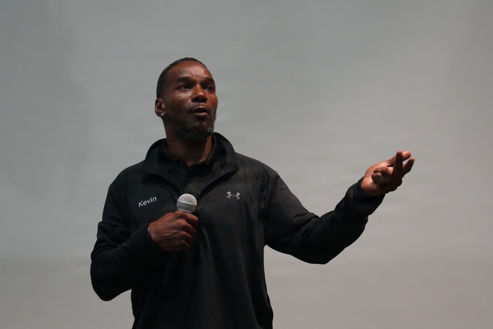 A man named Kevin, wearing a black Under Armour jacket with his name embroidered on the chest, is speaking into a handheld microphone. He appears to be giving a presentation or speech, gesturing with his right hand for emphasis. The background is a plain light gray surface, consistent with a stage or presentation setting.