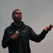 A man named Kevin, wearing a black Under Armour jacket with his name embroidered on the chest, is speaking into a handheld microphone. He appears to be giving a presentation or speech, gesturing with his right hand for emphasis. The background is a plain light gray surface, consistent with a stage or presentation setting.