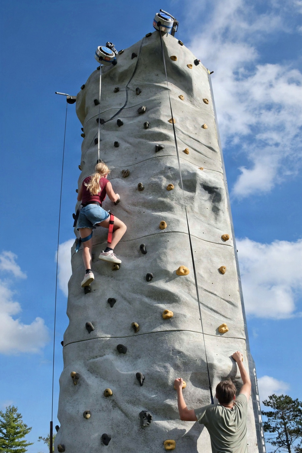 Two individuals are climbing an outdoor artificial rock climbing tower under a partly cloudy blue sky. The tower is tall and cylindrical, fitted with numerous colored hand and foot holds. One climber, a young woman with blonde hair, is approximately mid-way up the wall secured by a harness and rope, while a second person at the base is just beginning their ascent.
