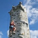 Two individuals are climbing an outdoor artificial rock climbing tower under a partly cloudy blue sky. The tower is tall and cylindrical, fitted with numerous colored hand and foot holds. One climber, a young woman with blonde hair, is approximately mid-way up the wall secured by a harness and rope, while a second person at the base is just beginning their ascent.
