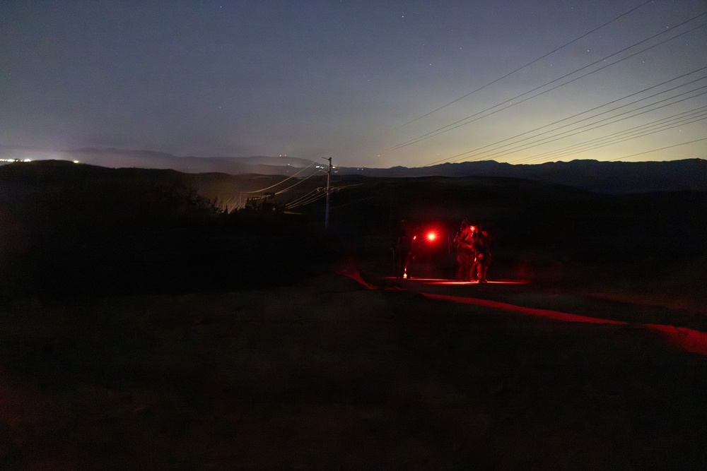A nighttime outdoor scene showing figures illuminated by red light alongside what appears to be a vehicle on a dirt road or path. The scene is set in a hilly or mountainous terrain with power lines visible in the background. City lights or ambient glow can be seen on the horizon, suggesting a populated area in the distance.