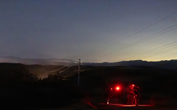 A nighttime outdoor scene showing figures illuminated by red light alongside what appears to be a vehicle on a dirt road or path. The scene is set in a hilly or mountainous terrain with power lines visible in the background. City lights or ambient glow can be seen on the horizon, suggesting a populated area in the distance.