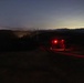 A nighttime outdoor scene showing figures illuminated by red light alongside what appears to be a vehicle on a dirt road or path. The scene is set in a hilly or mountainous terrain with power lines visible in the background. City lights or ambient glow can be seen on the horizon, suggesting a populated area in the distance.