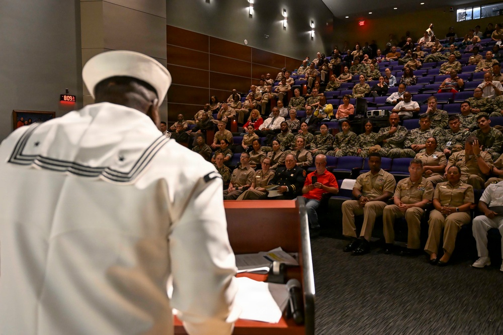 A U.S. Navy sailor in dress whites stands at a podium addressing a large auditorium filled predominantly with military personnel in tan/khaki uniforms. The setting appears to be a formal military briefing or ceremony held in a modern auditorium with purple seating. The audience includes a mix of Navy, Army, and possibly other branch personnel along with some civilians.