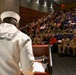 A U.S. Navy sailor in dress whites stands at a podium addressing a large auditorium filled predominantly with military personnel in tan/khaki uniforms. The setting appears to be a formal military briefing or ceremony held in a modern auditorium with purple seating. The audience includes a mix of Navy, Army, and possibly other branch personnel along with some civilians.