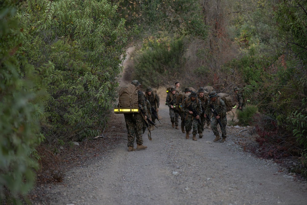 A group of military personnel in camouflage uniforms are conducting a foot march along a gravel trail through dense vegetation. They are carrying full combat loads including large rucksacks and rifles. The scene appears to be a training exercise based on the organized formation and safety reflective gear visible on some personnel.