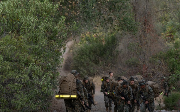 A group of military personnel in camouflage uniforms are conducting a foot march along a gravel trail through dense vegetation. They are carrying full combat loads including large rucksacks and rifles. The scene appears to be a training exercise based on the organized formation and safety reflective gear visible on some personnel.
