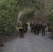 A group of military personnel in camouflage uniforms are conducting a foot march along a gravel trail through dense vegetation. They are carrying full combat loads including large rucksacks and rifles. The scene appears to be a training exercise based on the organized formation and safety reflective gear visible on some personnel.