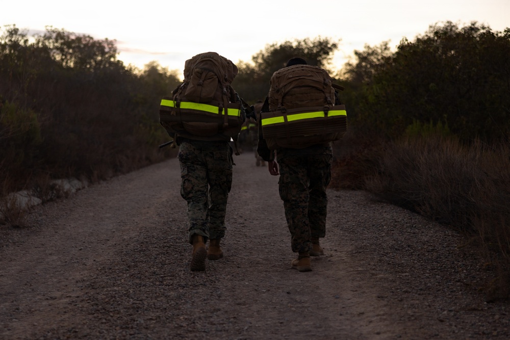 Two military personnel in camouflage uniforms are walking away from the camera along a gravel path during early morning or dusk conditions. Both carry large tactical backpacks equipped with high-visibility neon yellow reflective strips, suggesting a ruck march or training exercise. Additional personnel are faintly visible further down the trail.