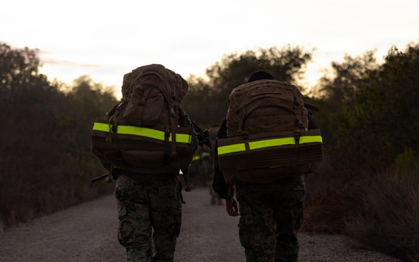 Two military personnel in camouflage uniforms are walking away from the camera along a gravel path during early morning or dusk conditions. Both carry large tactical backpacks equipped with high-visibility neon yellow reflective strips, suggesting a ruck march or training exercise. Additional personnel are faintly visible further down the trail.