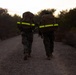 Two military personnel in camouflage uniforms are walking away from the camera along a gravel path during early morning or dusk conditions. Both carry large tactical backpacks equipped with high-visibility neon yellow reflective strips, suggesting a ruck march or training exercise. Additional personnel are faintly visible further down the trail.