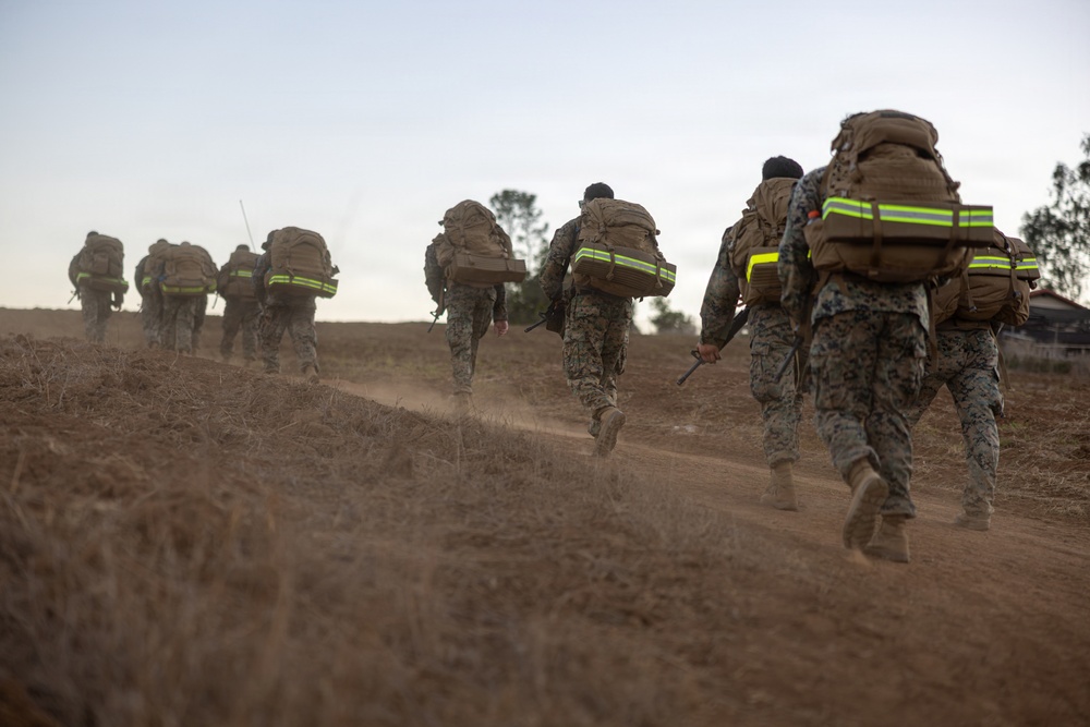 A column of military personnel in camouflage uniforms are conducting a ruck march along a dusty dirt trail in an arid, open landscape. All individuals are carrying large tactical backpacks equipped with high-visibility yellow-green reflective strips, suggesting a safety or training protocol. The image is taken from a low, rear-facing perspective during low-light conditions, likely dawn or dusk.