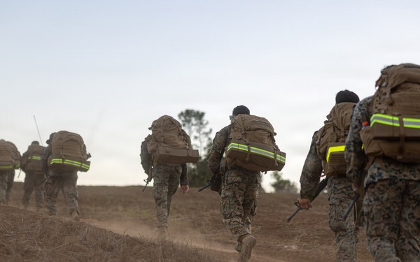 A column of military personnel in camouflage uniforms are conducting a ruck march along a dusty dirt trail in an arid, open landscape. All individuals are carrying large tactical backpacks equipped with high-visibility yellow-green reflective strips, suggesting a safety or training protocol. The image is taken from a low, rear-facing perspective during low-light conditions, likely dawn or dusk.