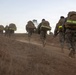 A column of military personnel in camouflage uniforms are conducting a ruck march along a dusty dirt trail in an arid, open landscape. All individuals are carrying large tactical backpacks equipped with high-visibility yellow-green reflective strips, suggesting a safety or training protocol. The image is taken from a low, rear-facing perspective during low-light conditions, likely dawn or dusk.